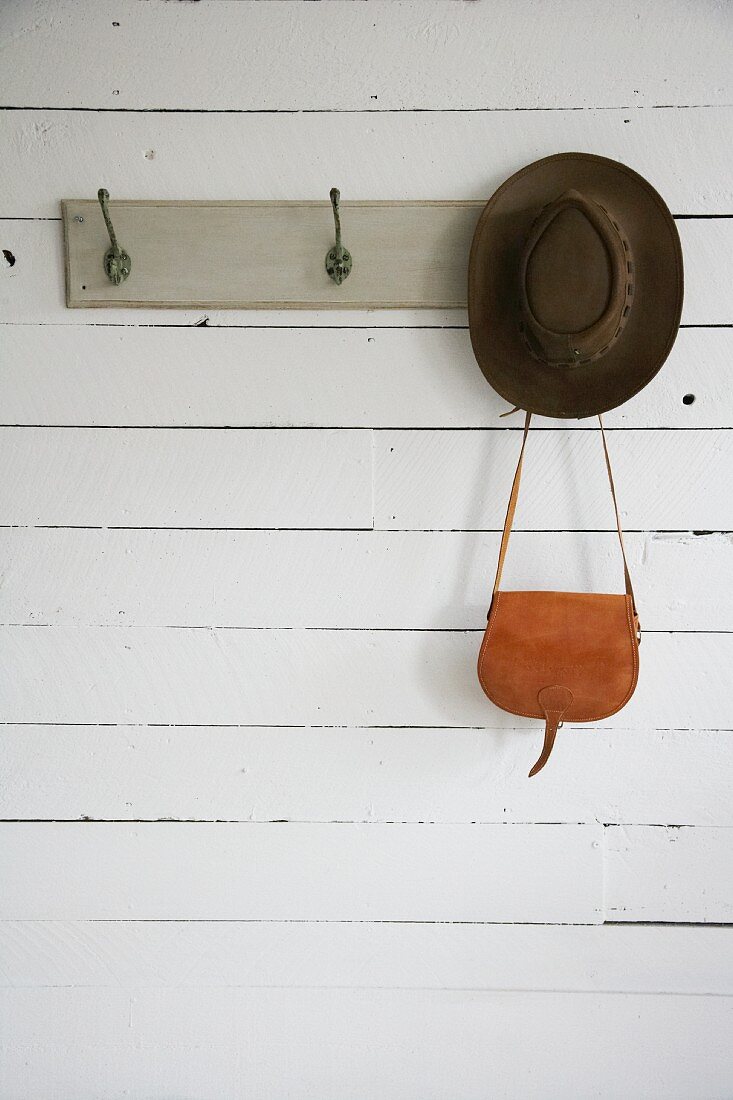 Hat and leather bag hanging from coat rack painted grey on white board wall