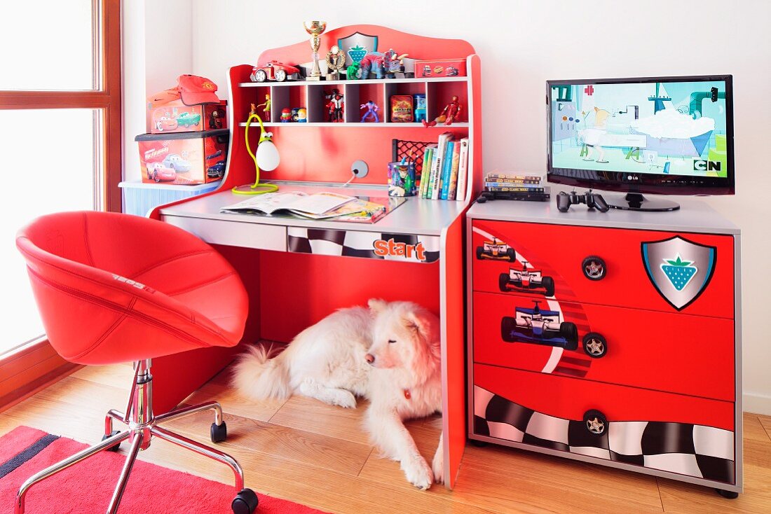 Roter Schreibtisch mit Regalaufsatz und Kommode mit aufgedruckten Rennwagen-Symbolen; Hund unter dem Tisch