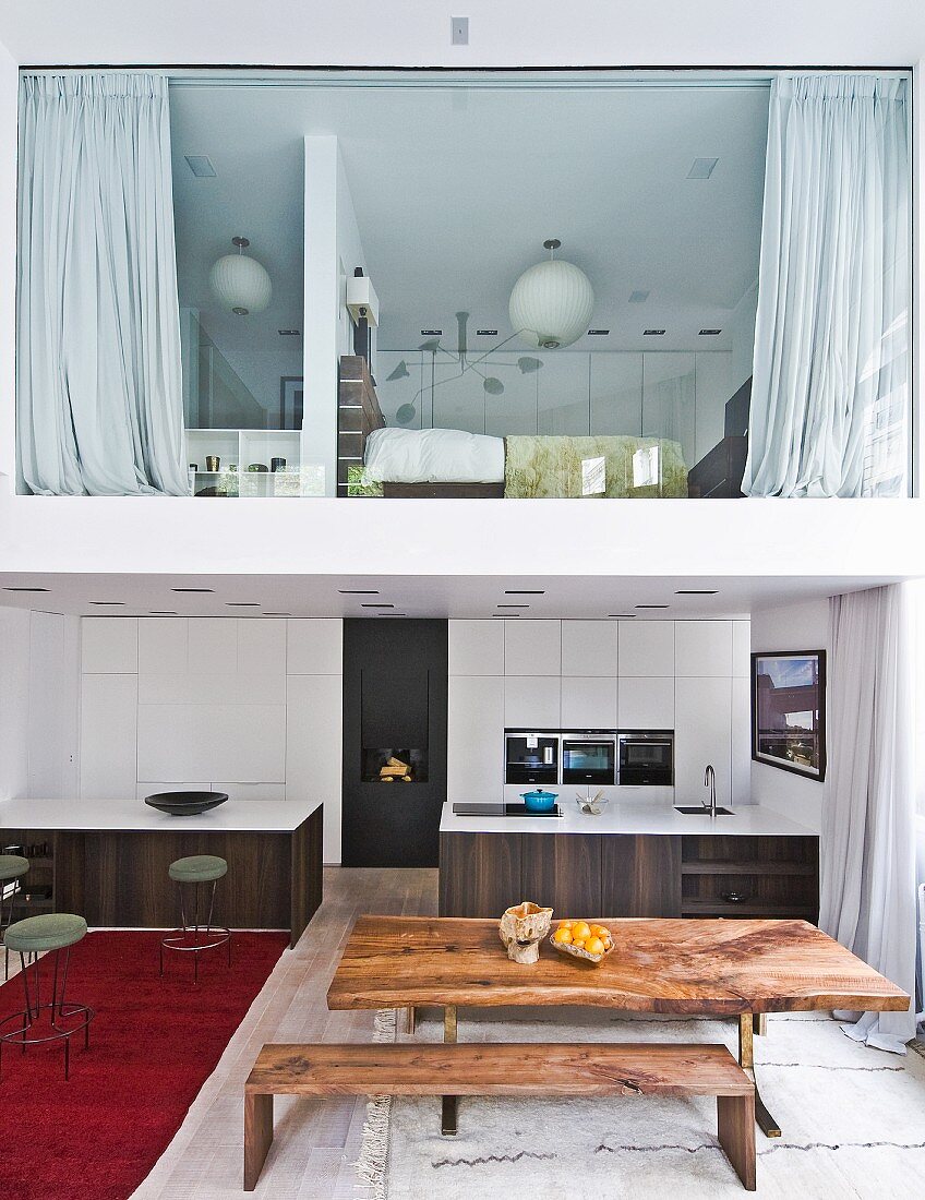 Wooden table and matching bench in front of open-plan kitchen with two monolithic counters below mezzanine with glass wall