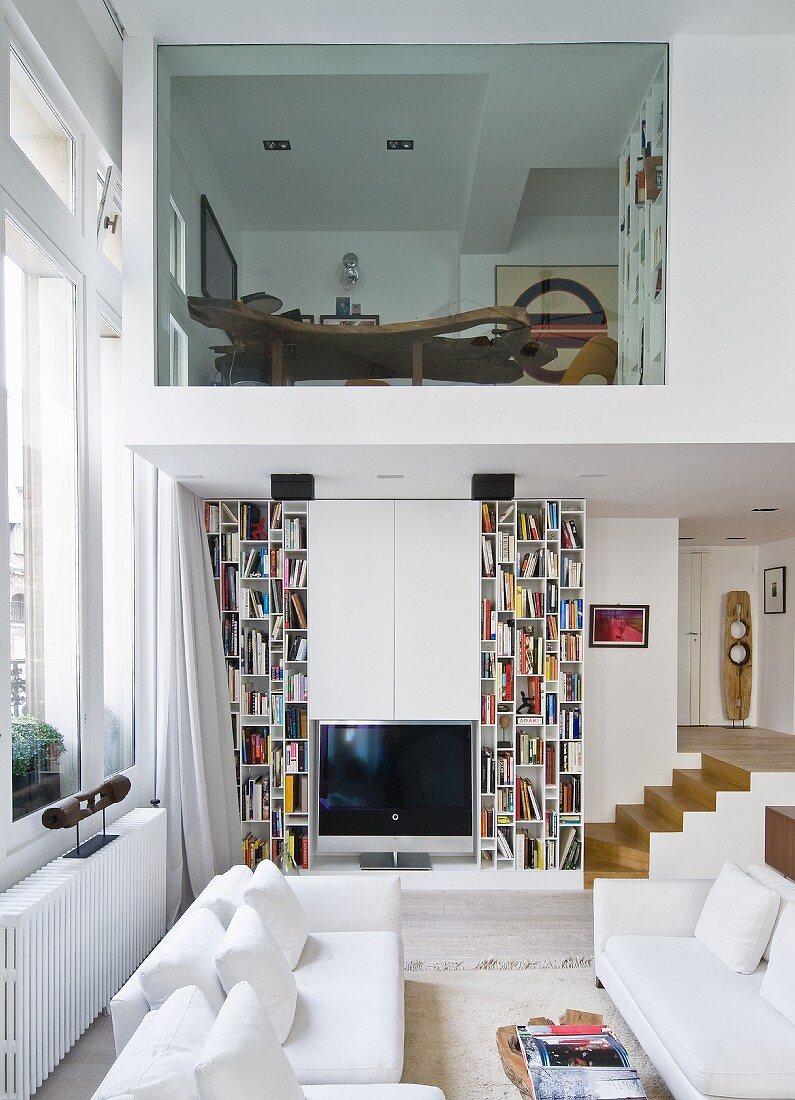 White sofa set and designer bookcases in living room, partly double-height; view of office in upper storey with glass wall