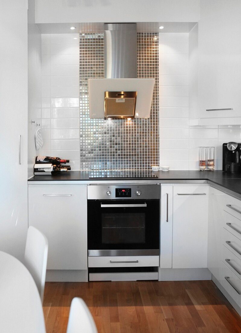Modern, white fitted kitchen with stainless steel extractor hood and splashback of reflective mosaic tiles