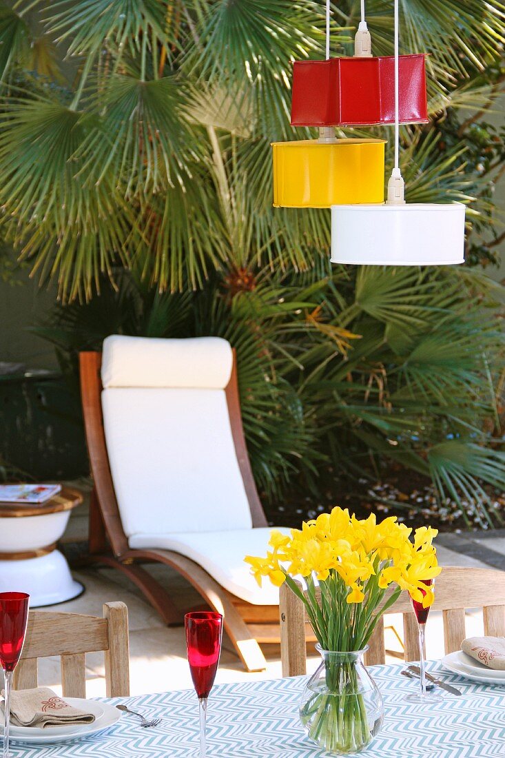 Ehemalige Backformen in verschiedenen Farben als Hängeleuchten über Terrassentisch, Holz Liegestuhl mit weissen Polstern vor Palme