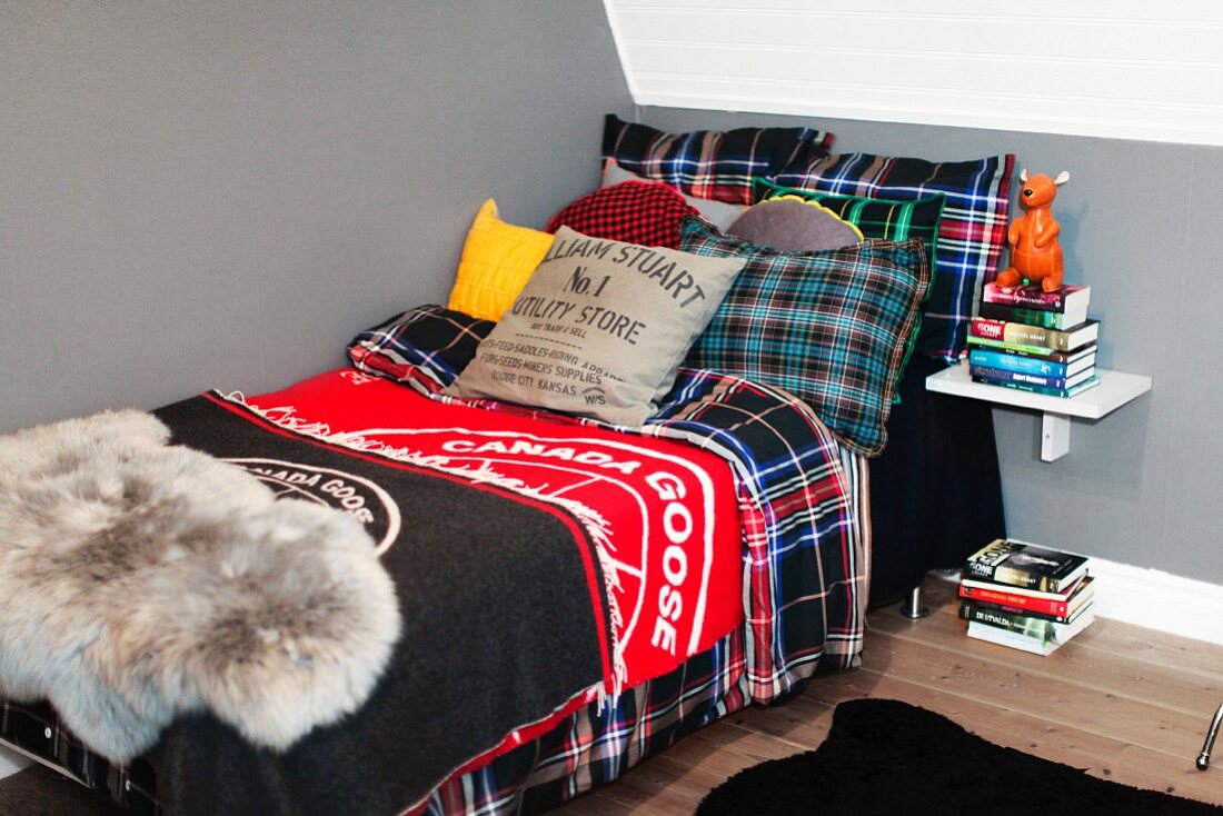 Cosy teenager's bedroom with single bed, multiple tartan scatter cushions and bedspread and stacked books on floor and on wall bracket
