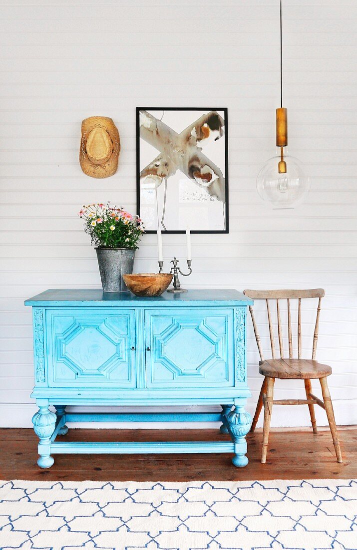 Pale blue, carved dresser and country-house wooden chair below modern artwork on white-painted, wood-clad wall and retro pendant lamp