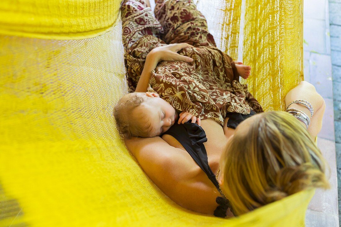 Junge Mutter mit schlafendem Baby auf dem Bauch in einer gelben Hängematte