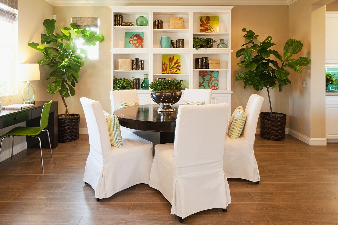 Wohnraum mit rundem Esstisch, Hussenstühlen & von Grünpflanzen eingerahmtem Regalschrank