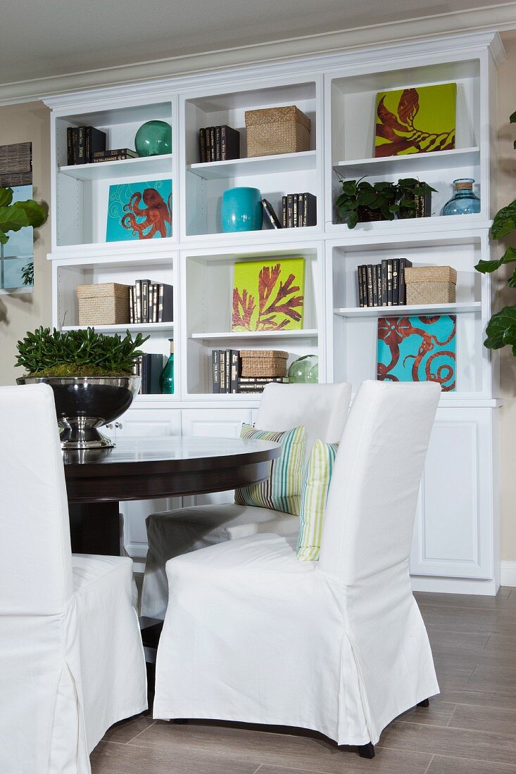 weiße Hussenstühle an rundem Esstisch vor weißem Regalschrank mit Büchern & Dekoobjekten