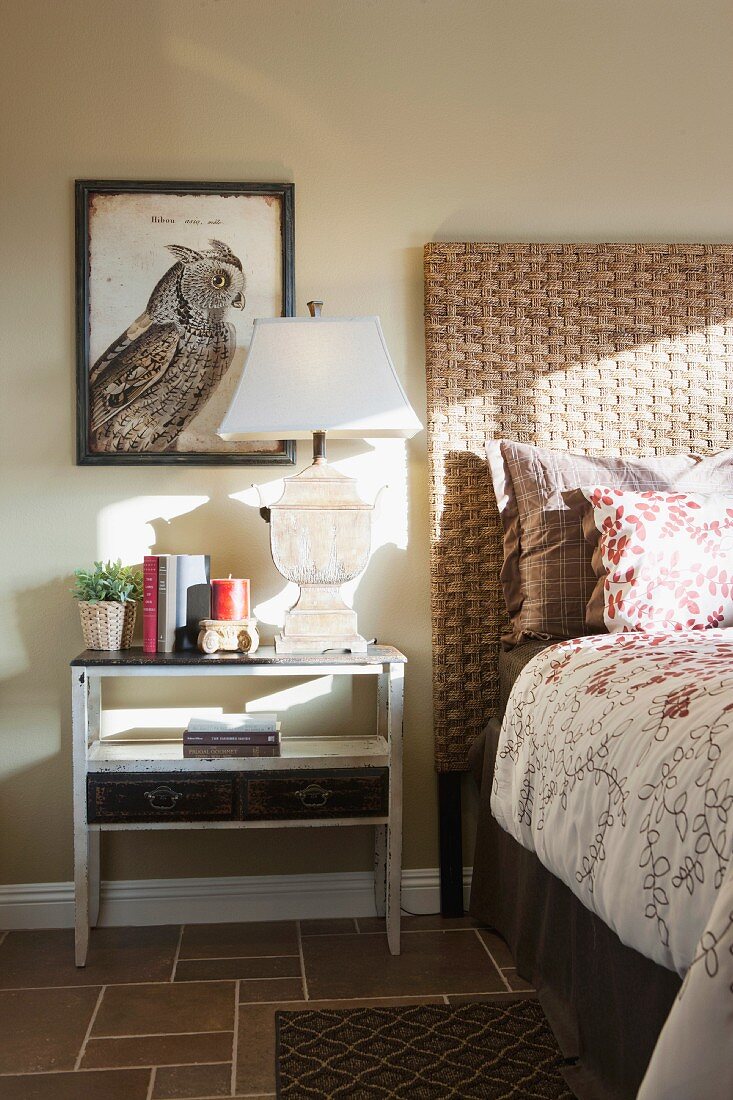 Schlafzimmer mit Nachttisch, Tischleuchte & Wandbild mit Vogelmotiv neben Bett mit geflochtenem Kopfteil