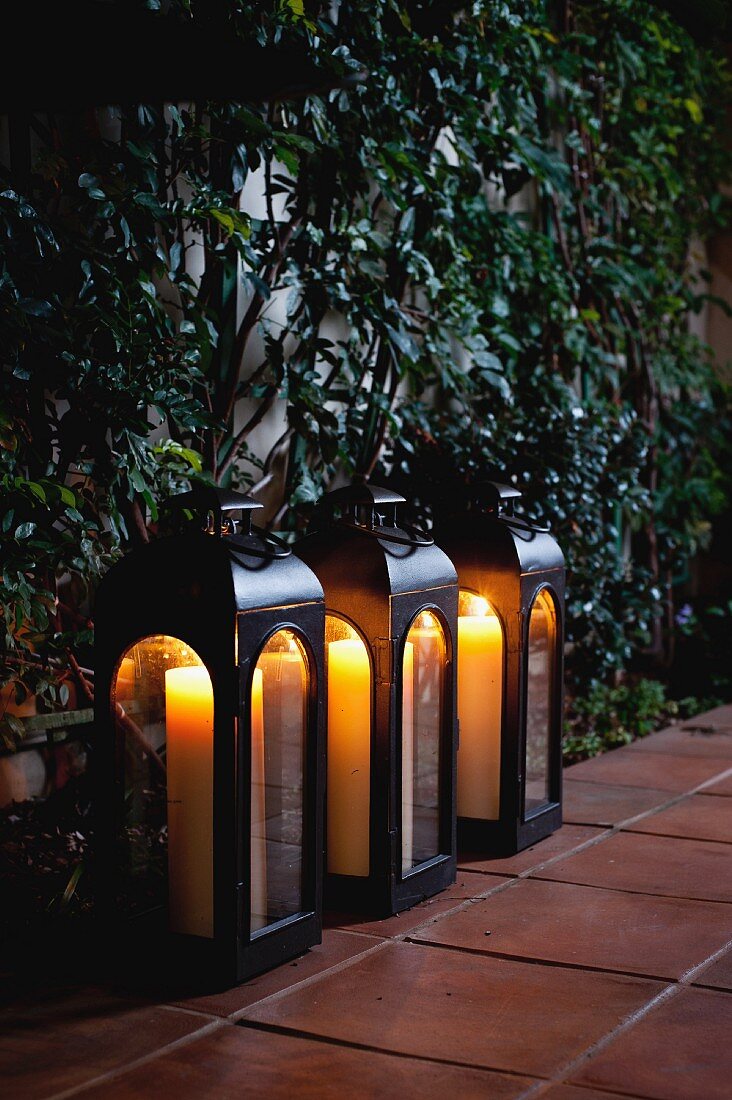 Drei leuchtende Kerzenlaternen auf Gehweg vor berankter Hauswand
