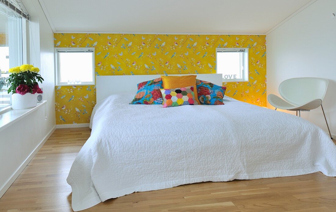 Doppelbett mit Kopfteil, weisser Tagesdecke und bunten Kissen vor gelber Tapete mit Vogelmotiven, seitlich weißer Designerstuhl