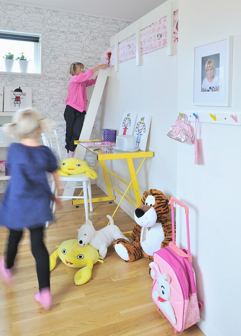 Spielende Mädchen im Kinderzimmer mit Bettleiter zur Schlafebene, gelbem Schreibtisch und Stofftieren am Boden