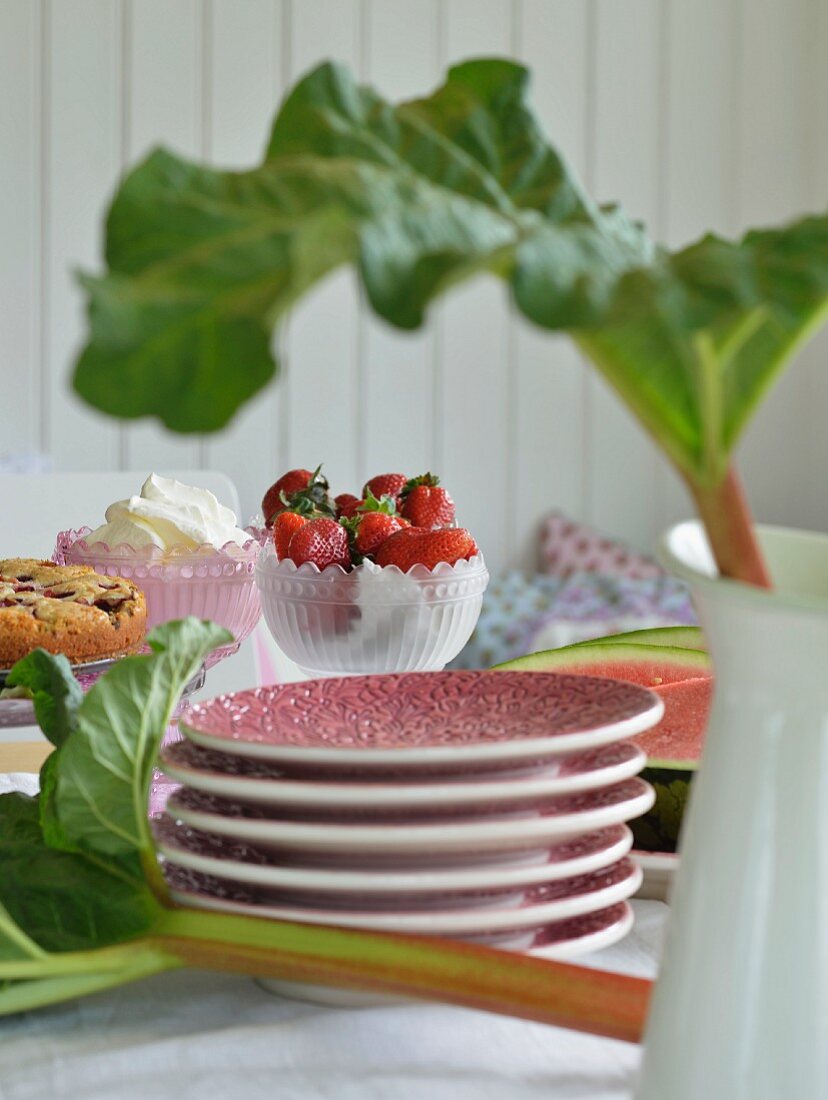 Stacked plates amongst rhubarb leaves, cakes and bowl of strawberries on table set for afternoon tea