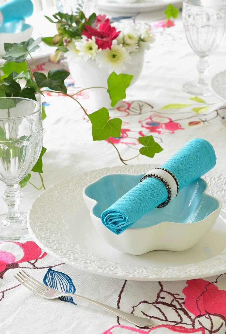 Türkisblaue Stoffserviette mit Serviettenring auf weisser Schale auf floraler Tischdecke