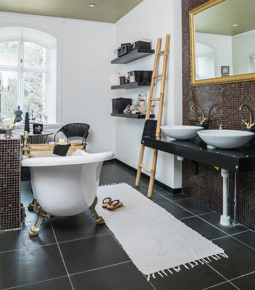 Antique, free-standing bathtub, washstand and gilt-framed mirror in black and white bathroom with ornaments and accessories