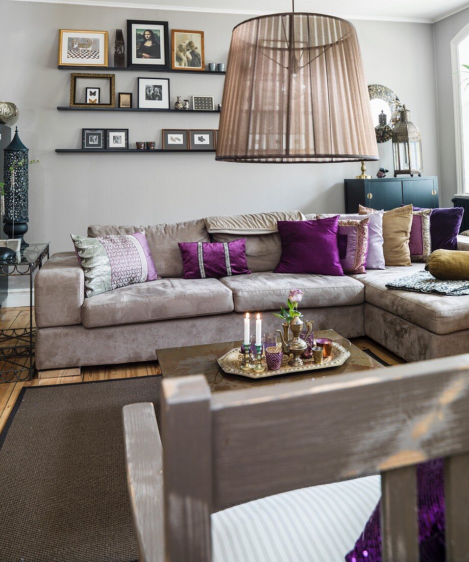 Sand-coloured sofa with purple scatter cushions below floating shelves on grey-painted wall
