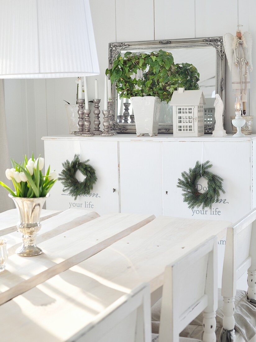 weiße Tulpen in Silberkelch auf rustikaler, weisser Tischplatte; mit Kerzen, Pflanze & Spiegel dekoriertes Sideboard im Hintergrund