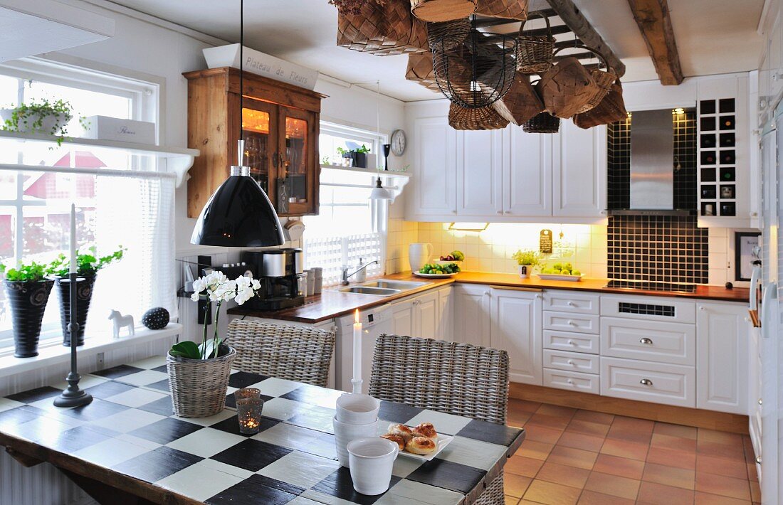 Table with painted chequered top and wicker chairs in country-house kitchen-dining room with baskets hanging from ceiling