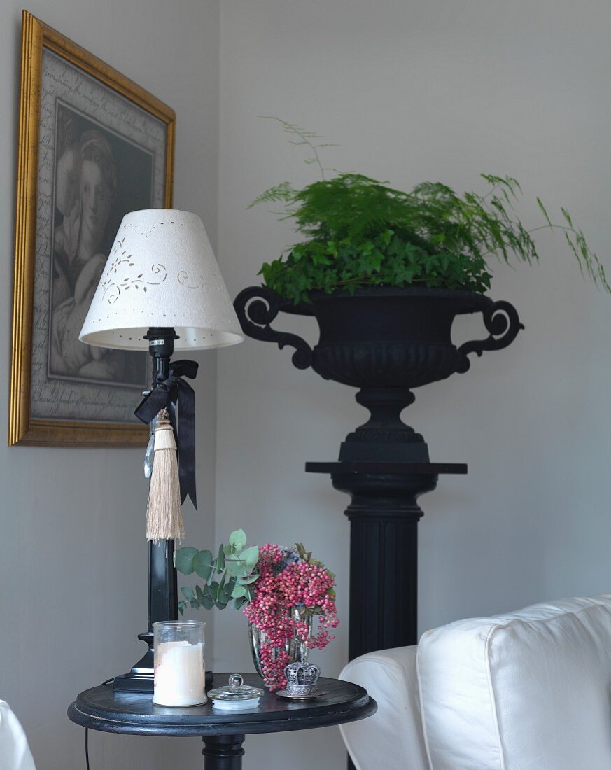 Table lamp with white lampshade on round side table, and foliage plants in black, antique urn on plinth in background