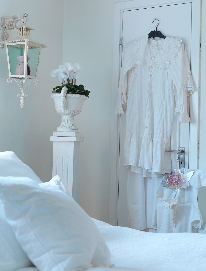Blick über Bett auf Stele mit antikem Pflanzengefäss neben Tür, darauf gehängtes Nachthemd auf Kleiderbügel
