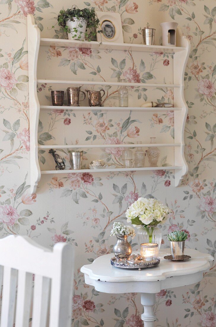 Vase of flowers on side table below shelving on wall with floral wallpaper