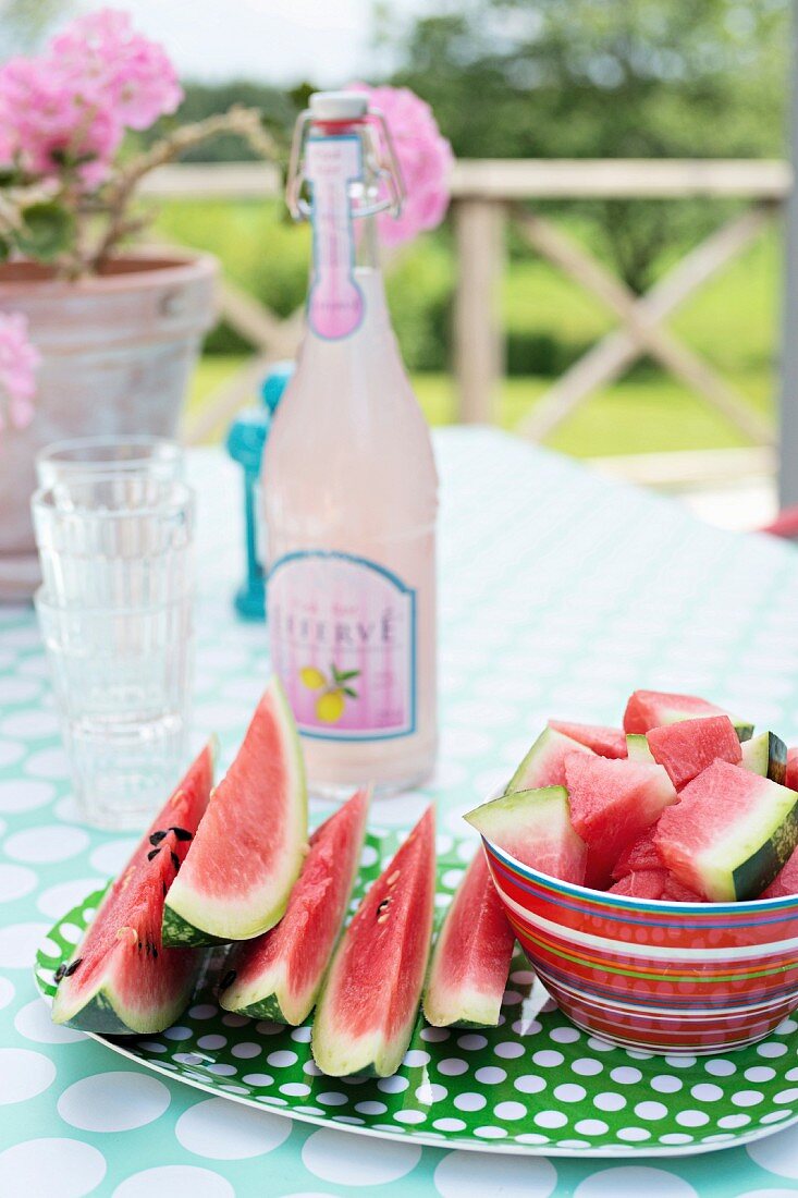 Grün-weiß gepunkteter Teller und rote Streifenschale mit geschnittener Wassermelone dahinter Retro Limonadenflasche auf sommerlichem Gartentisch