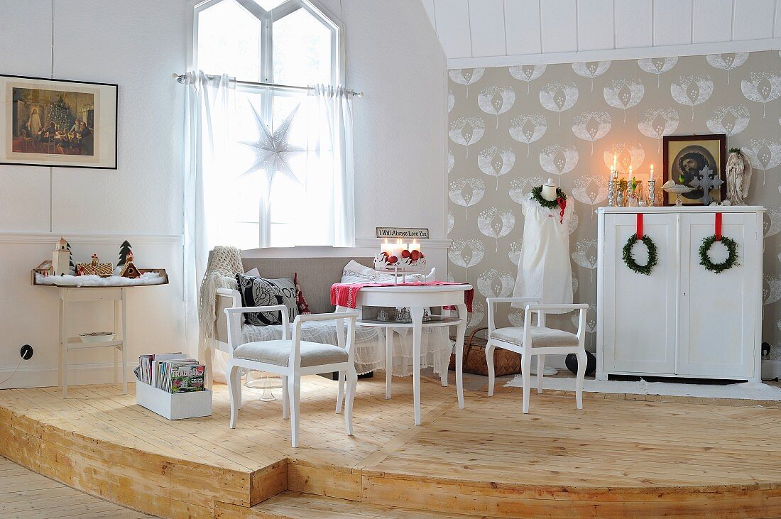 Romantic, white, shabby-chic sitting area on curved wooden dais; festive wreaths hung from red ribbons