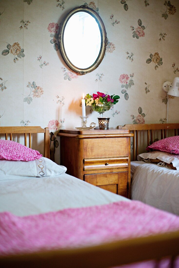 Wood-framed twin beds and matching bedside cabinet against floral wallpaper in rustic bedroom