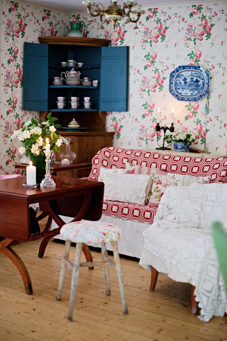 Verschiedene Sitzmöbel neben antikem Holztisch und Sofa, seitlich offener Eckschrank mit Geschirr vor Wand mit geblümter Tapete
