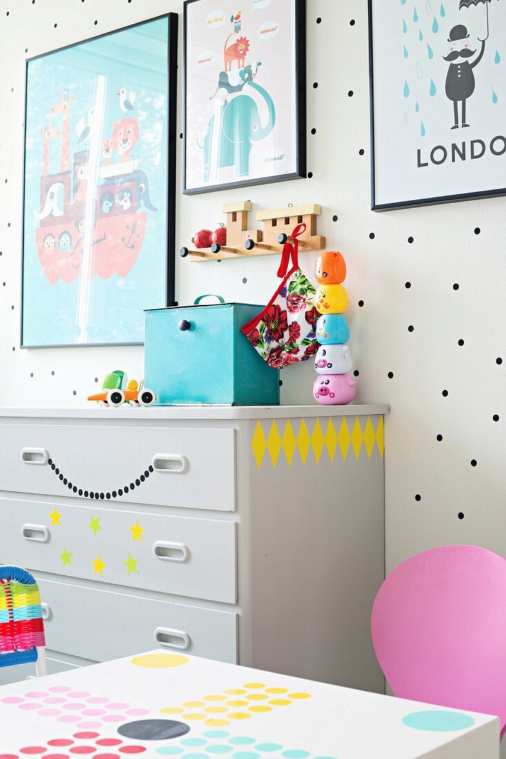 Gerahmte Plakate an weißer Wand mit schwarzen Punkten und Kinderspielzeug auf grauer Kommode mit Deko-Aufklebern