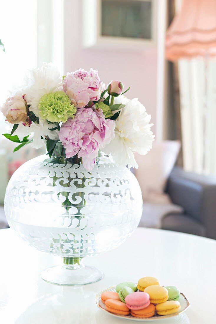 Blumenstrauss in Glasvase mit geätztem Muster und bunte Macarons auf weißem Tisch
