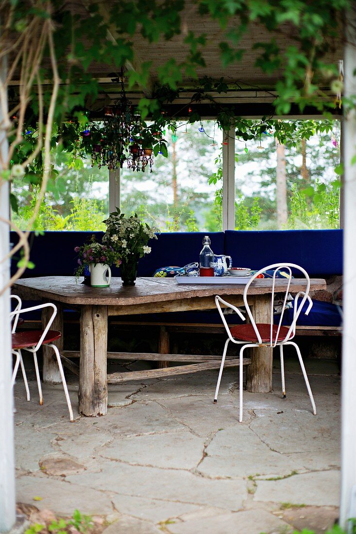 Rustikaler Holztisch und weiße, filigrane Metallstühle auf Natursteinboden in Loggia