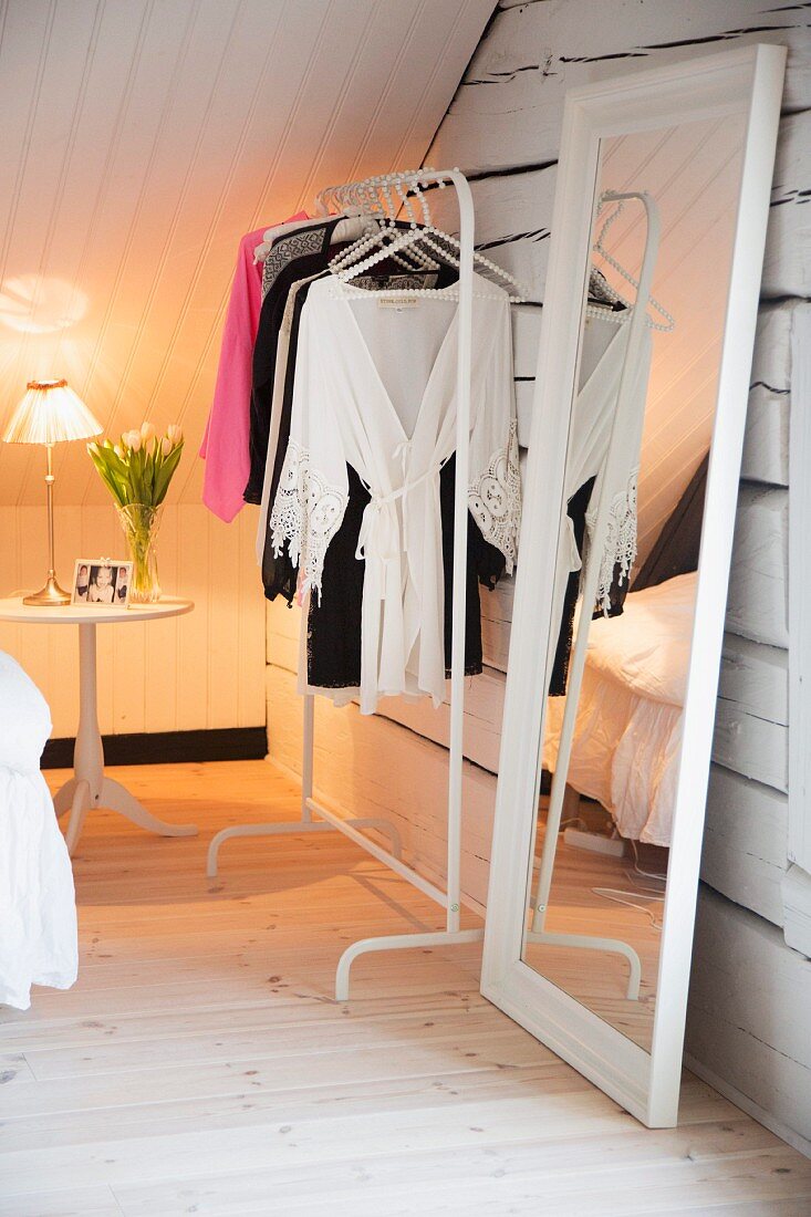 White, feminine attic room with full-length mirror, clothes rack and tulips on bedside table in background
