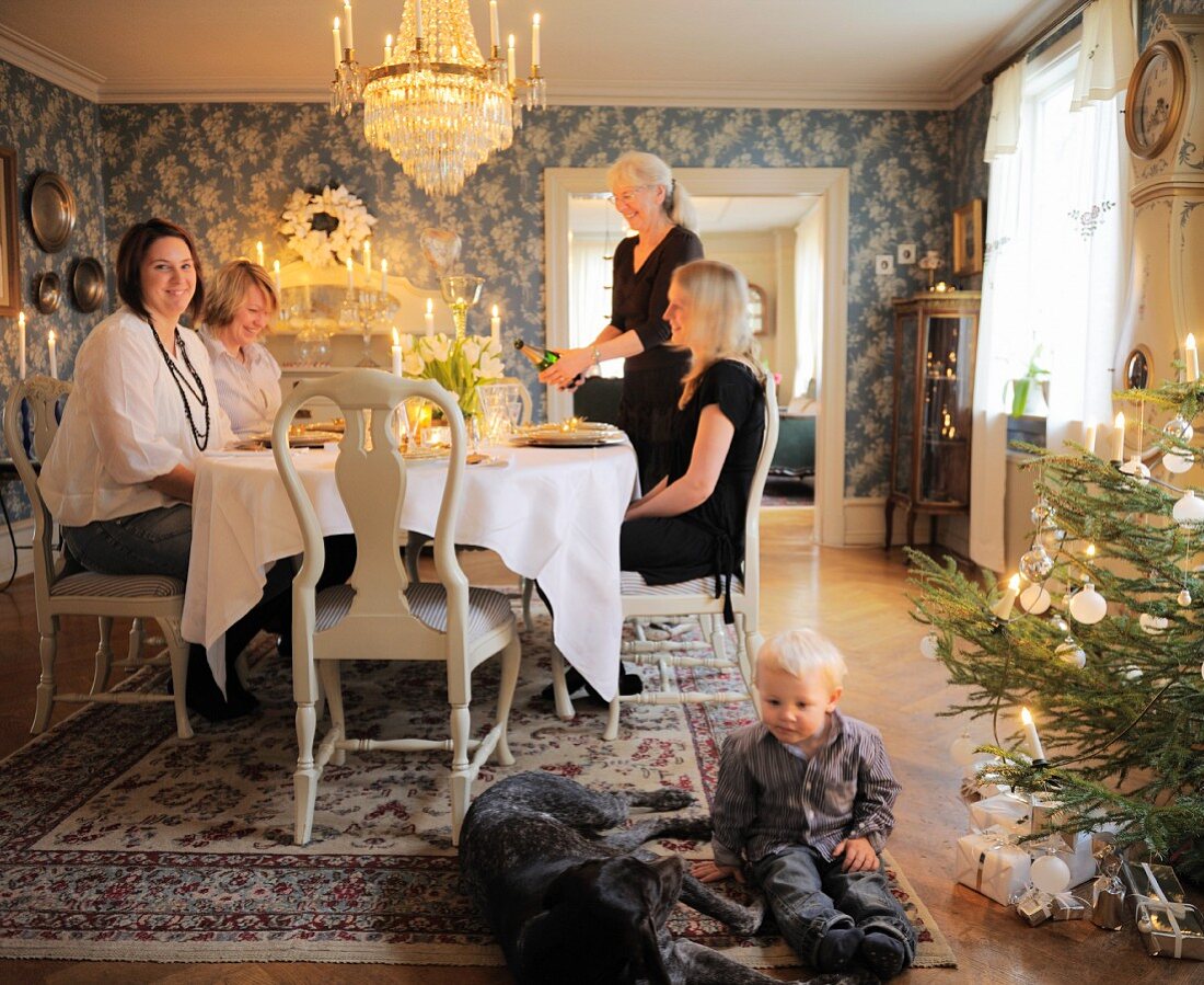 Toddler and dog sitting on rug next to Christmas tree in front of family around dinner table in traditional dining room