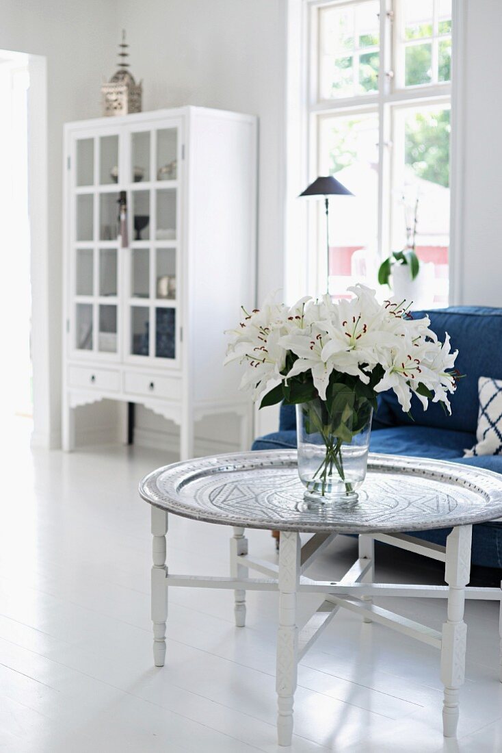 Weißer Lilienstrauss auf Tabletttisch, blaues Polstersofa und weisser Vitrinenschrank im Hintergrund