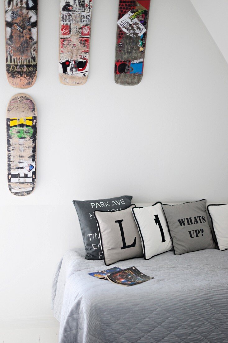 Scatter cushions printed with letters on grey throw on teenager's bed below scuffed skateboards decorating wall