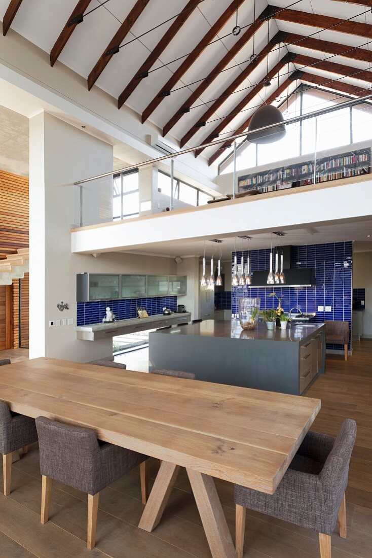 Pale wooden dining table and chairs with grey upholstery in front of kitchen island; mezzanine level and view of exposed roof structure in open-plan country house