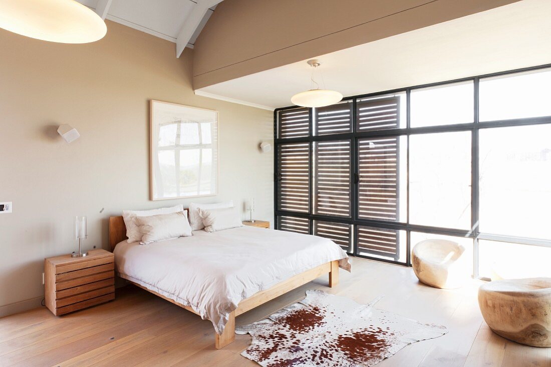 Kuhfell auf edlem Dielenboden vor schlichtem Doppelbett in grosszügigem Schlafzimmer, an Fensterfront Schiebeelemente aus Holzlamellen