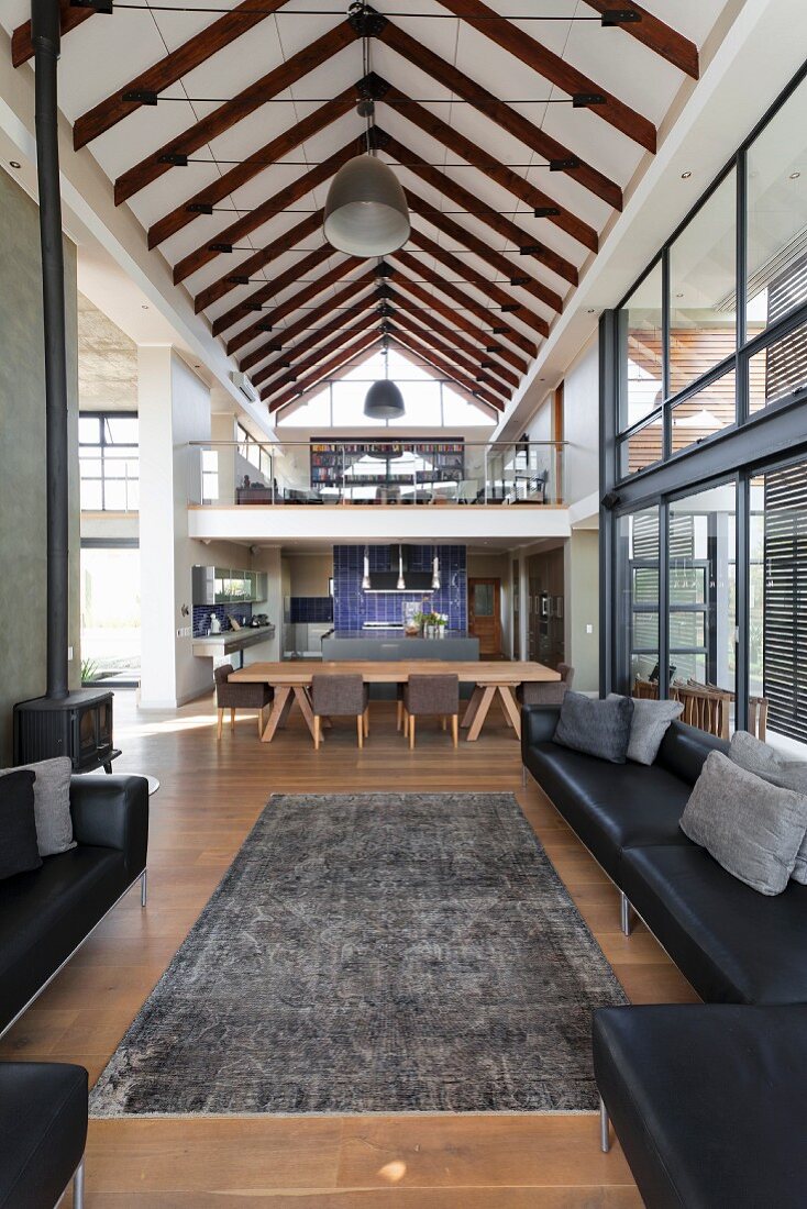 View from lounge area with black leather sofa set and vintage rug to dining area and mezzanine below exposed roof structure in contemporary home