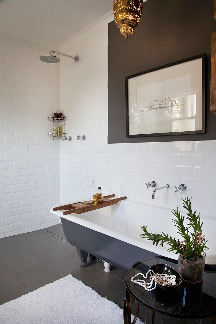 Plant on side table next to free-standing bathtub, framed drawing on wall and floor-level, open shower in corner of white-tiled bathroom