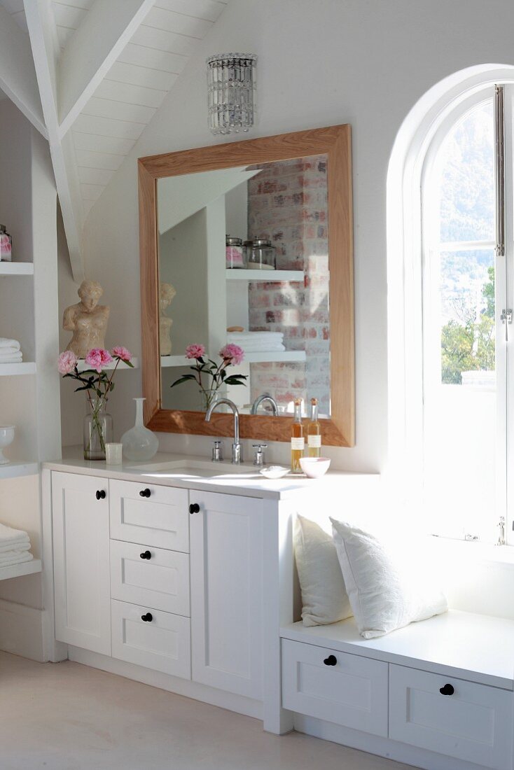 Bathroom with white washstand and bench below framed mirror and arched window