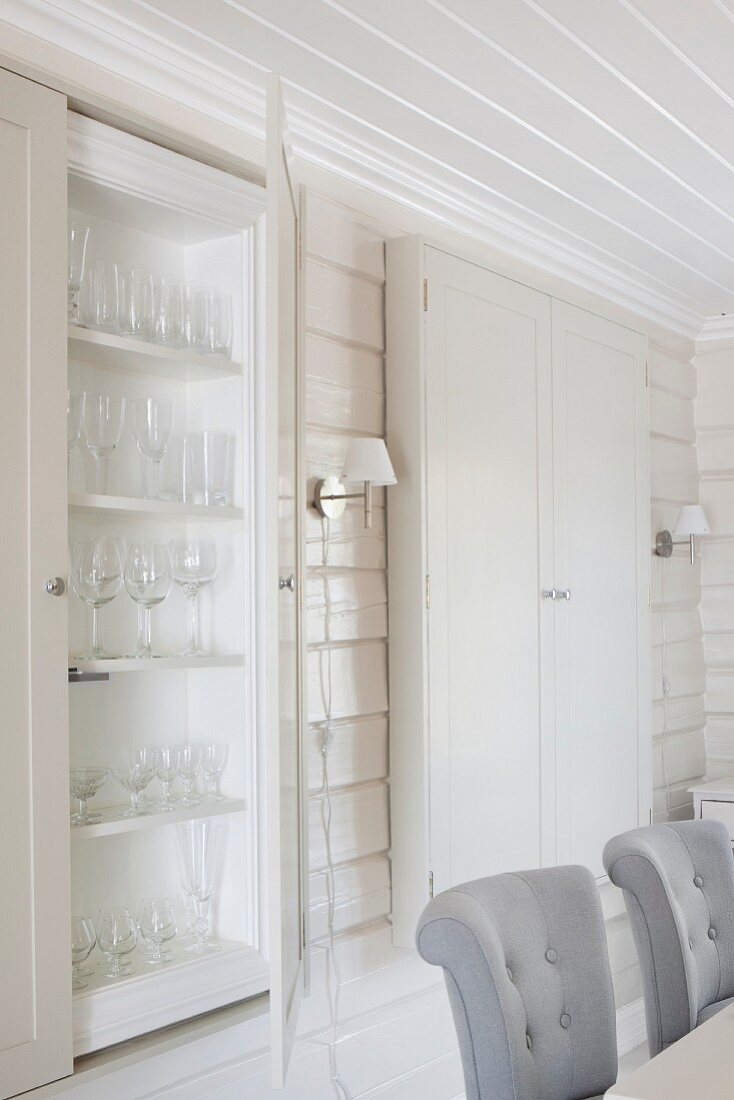 Backrests of upholstered dining chairs and fitted display cabinets in interior with white, wood-clad walls and ceiling