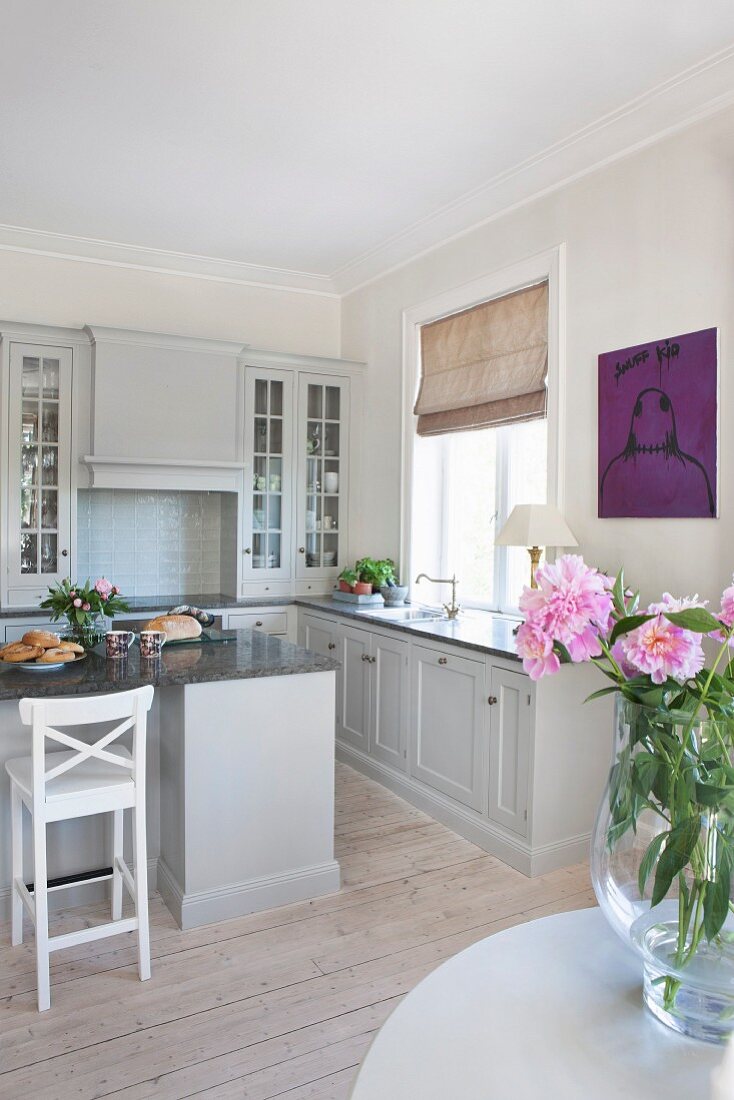 Offene Küche mit freistehendem Mittelblock, helle Fronten im Landhausstil, im Vordergrund Blumenvase auf Tisch