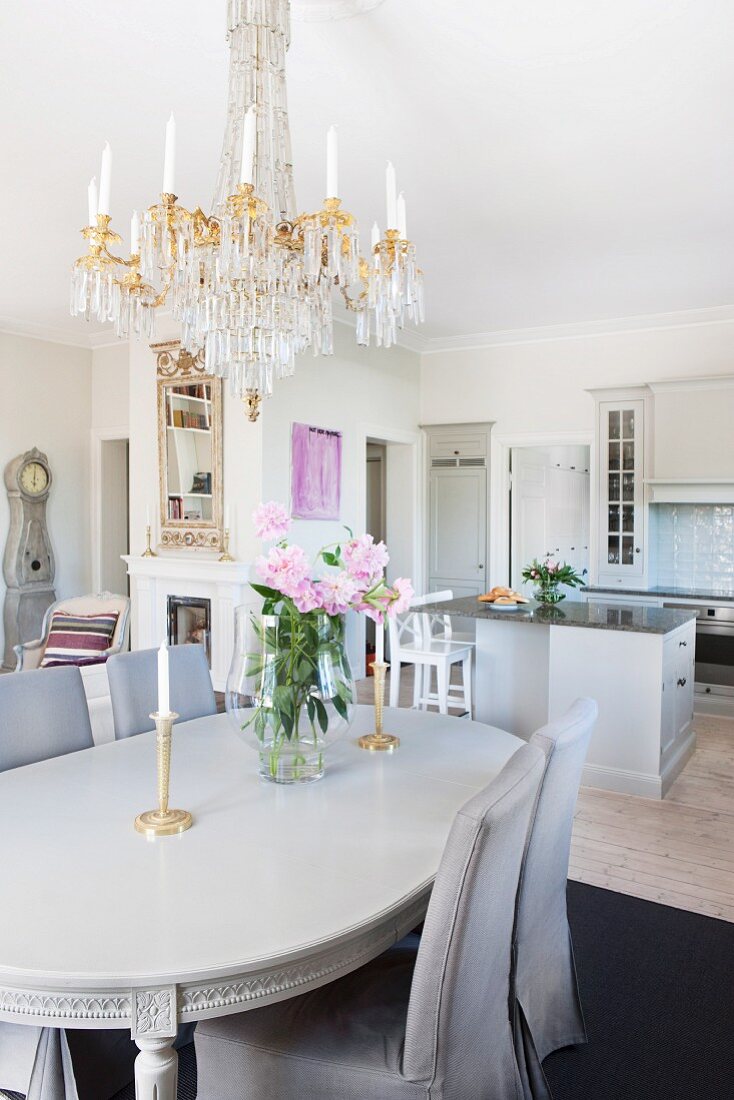 Stühle mit grauen Hussen um klassizistischen Esstisch unter Kronleuchter in offenem Wohnraum mit Küche