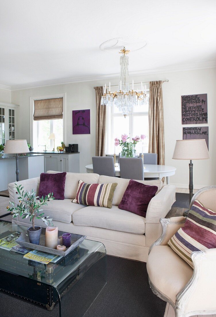 Helles Sofa und Sessel mit gestreiftem Kissen um Couchtisch aus gebogenem Glas in offenem Wohnraum