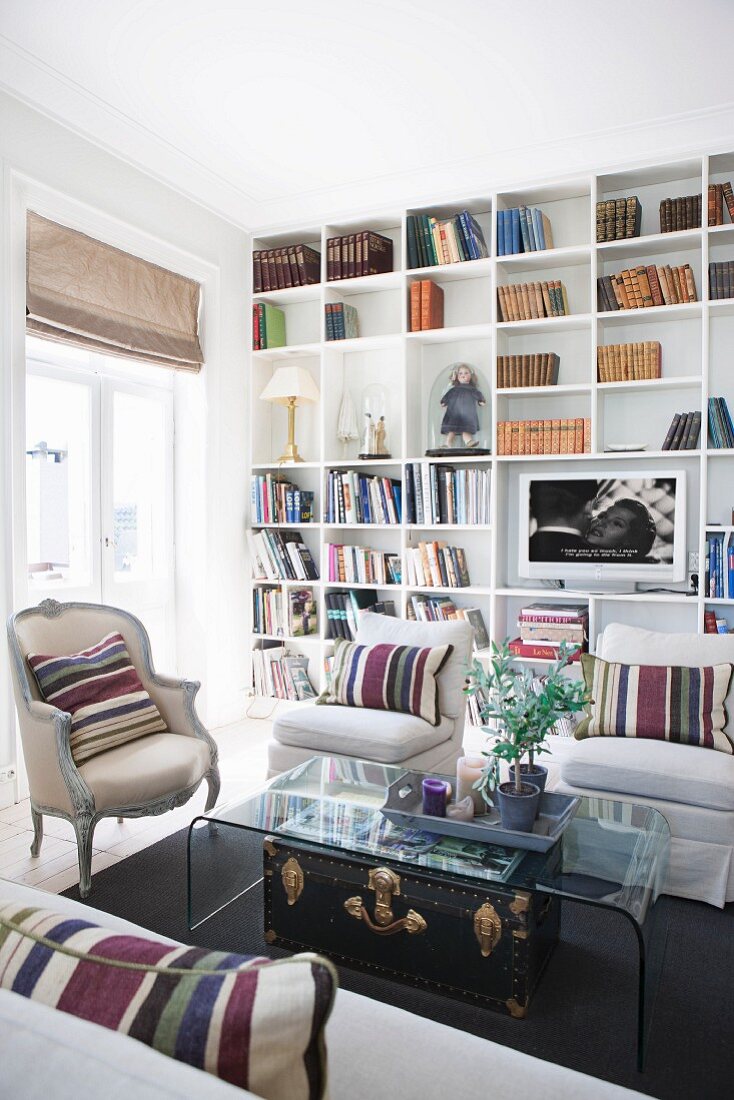 Striped scatter cushions on pale armchairs and Rococo armchair around curved glass table in front of fitted bookcases