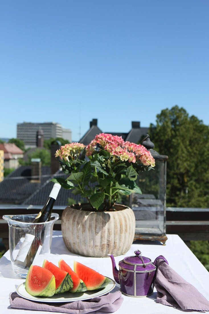 Melonenstücke auf Teller und Blumentopf mit Hortensien auf Tisch mit weisser Tischdecke auf dem Balkon
