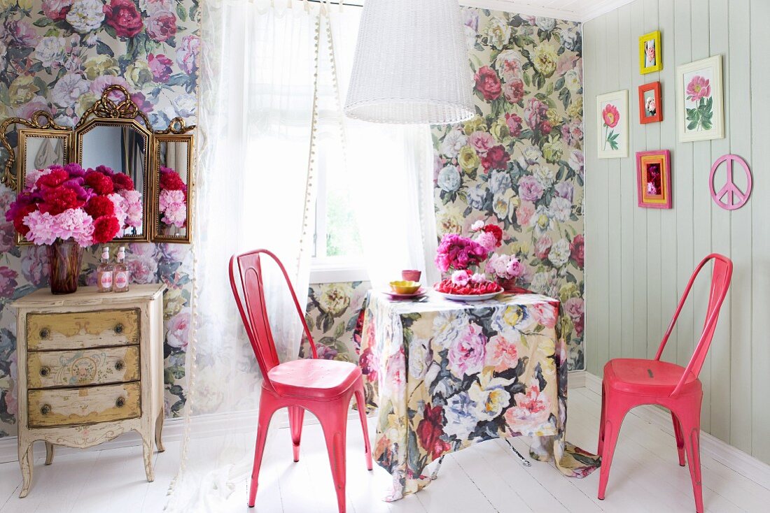 Blumentapete, passende Tischdecke und pinkfarbene Retro-Metallstühle; seitlich Vintage Kommode mit Pfingstrosen und Zierspiegel