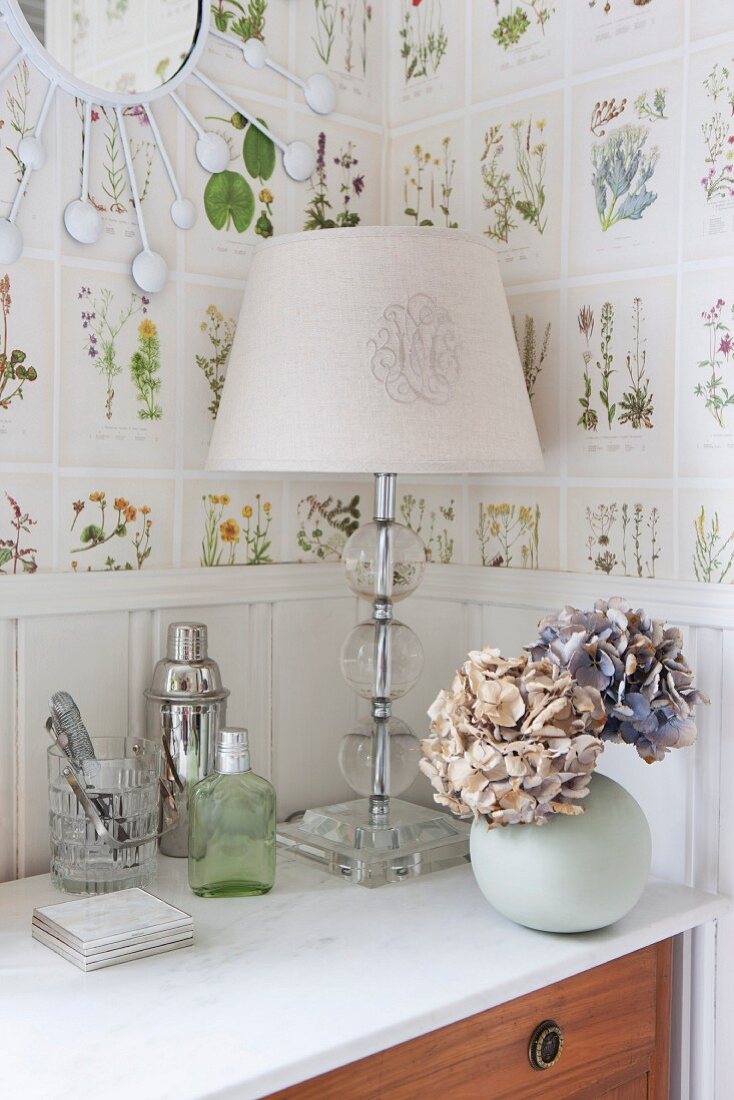 Tischleuchte mit weißem Schirm und Hortensien in Kugelvase auf Kommode, an Wand Tapete mit botanischem Muster