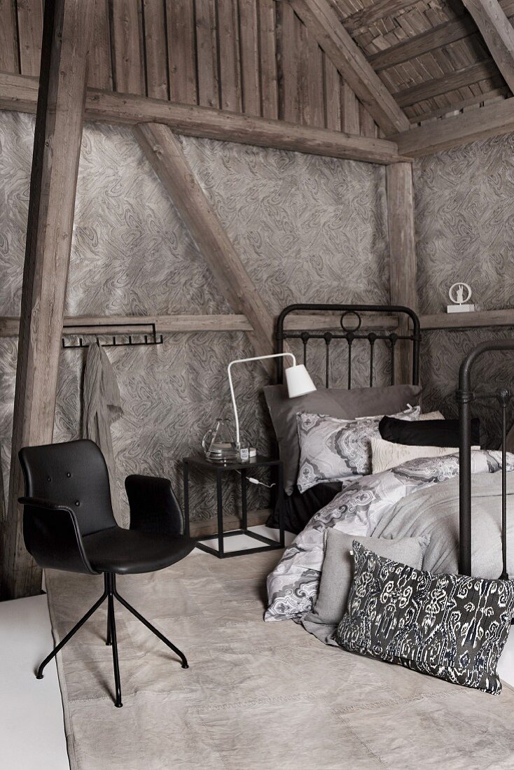 Bedroom in various shades of grey in barn-style interior with armchair, metal bed and half-timbered walls with wallpaper panels