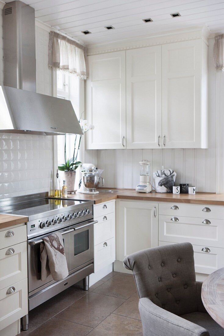 White country-house kitchen with stainless steel cooker and grey upholstered chair in foreground